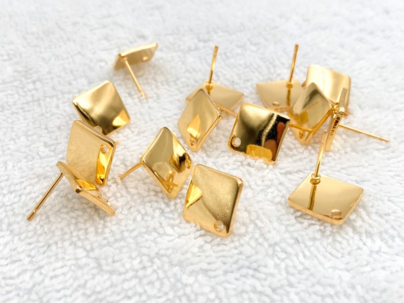 May include: A group of gold-toned square earring blanks with a small hole in the centre of each square. The earring blanks are scattered on a white surface.