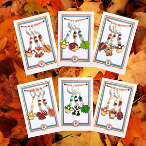 May include: Six sets of colorful stitch markers featuring woodland animal charms. Each set includes a mushroom, a leaf, and an animal charm, such as a fox, hedgehog, deer, squirrel, badger, and owl. The charms are attached to silver rings with colorful beads.