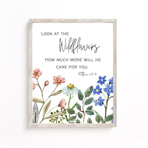 Könnte beinhalten: Ein weißer gerahmter Druck mit einer Aquarell-Illustration von Wildblumen. Der Druck zeigt den Text "Schau dir die Wildblumen an. Wie viel mehr wird er sich um dich kümmern. Matthäus 6,28-30"