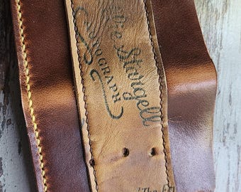 Peut inclure: Un portefeuille en cuir marron fait à la main avec des coutures jaunes. Le portefeuille présente un accent en cuir de gant de baseball vintage avec le texte "The Field" et "De Strugell" imprimé.