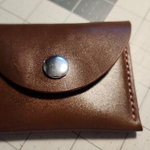 May include: A small, brown leather wallet with a flap secured by a silver snap. The edges are stitched with pink thread, adding a contrasting detail. The wallet is likely designed to hold cards or small items.