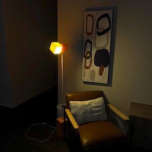 May include: A brown leather armchair with a white cushion sits in a dimly lit room. A floor lamp with a square orange shade illuminates the chair. A framed abstract painting with geometric shapes in brown, orange, and blue hangs on the wall.