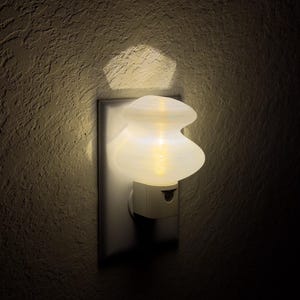 Puede incluir: Una luz nocturna de cerámica blanca enchufada a una toma de corriente. La luz nocturna tiene la forma de un objeto redondeado, bulboso, de dos niveles. La luz está encendida y brillando.