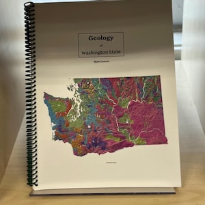 May include: A spiral-bound book titled "Geology of Washington State" by Ryan Lennon. The cover features a colorful geological map of Washington State, with shades of pink, green, and blue. The book is resting on a wooden shelf.