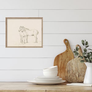 May include: A framed black and white pencil drawing of two horses standing side by side. The drawing is on a cream-colored background and is framed in a light wood frame.