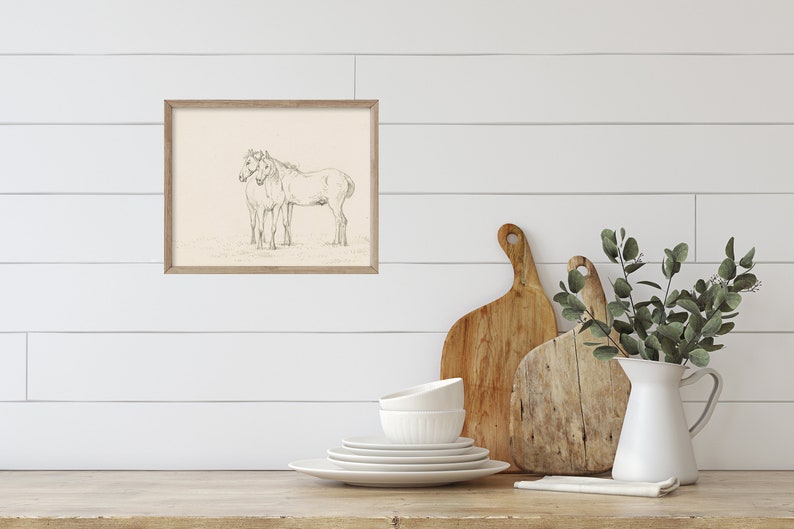 May include: A framed black and white pencil drawing of two horses standing side by side. The drawing is on a cream-colored background and is framed in a light wood frame.
