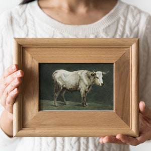 May include: A framed painting of a white cow standing in a field. The frame is made of wood and has a simple design.