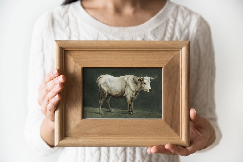 May include: A framed painting of a white cow standing in a field. The frame is made of wood and has a simple design.