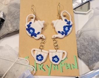 May include: A pair of dangle earrings featuring a white teapot and a white teacup with blue floral accents. The teapot and teacup are connected by a gold chain.