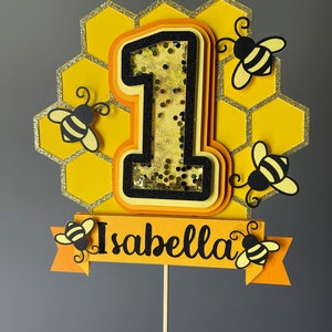 May include: A yellow and black bee-themed cake topper featuring the number "1" at the centre, set against a honeycomb backdrop. The number is filled with glitter. The name "Isabella" is displayed on a banner with bee accents. The topper is attached to a wooden stick.