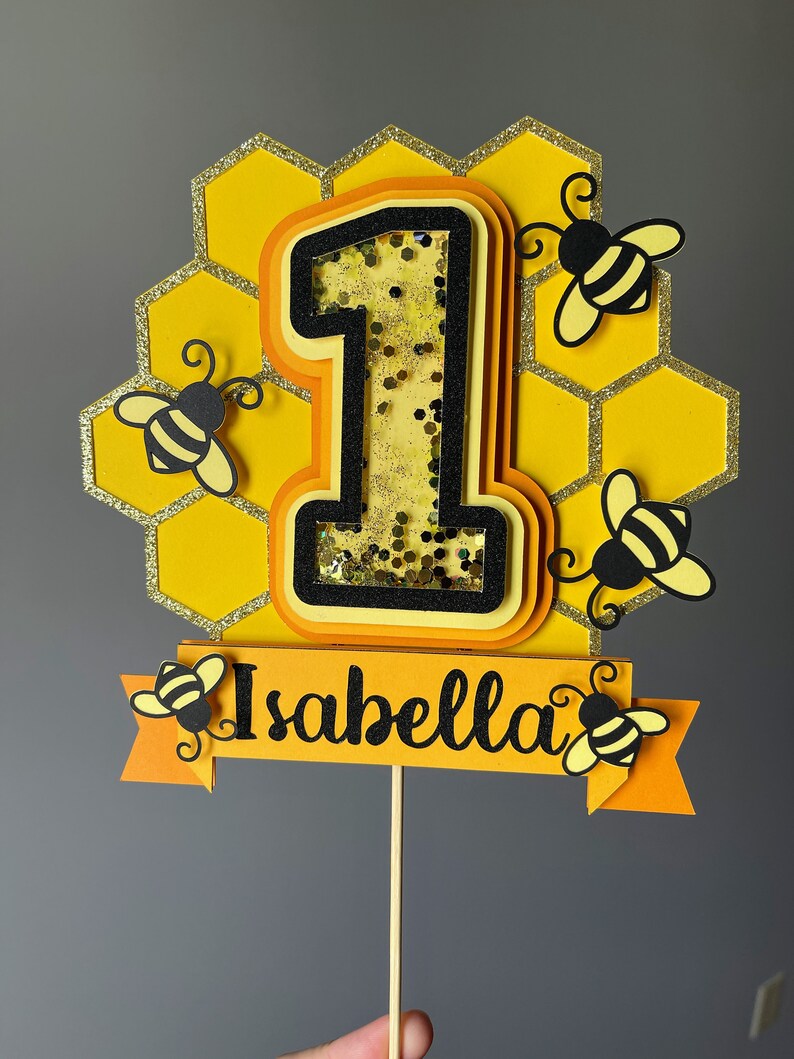 May include: A yellow and black bee-themed cake topper featuring the number "1" at the centre, set against a honeycomb backdrop. The number is filled with glitter. The name "Isabella" is displayed on a banner with bee accents. The topper is attached to a wooden stick.