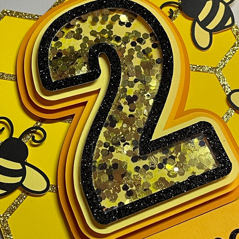 May include: A layered cake topper featuring the number two in yellow and black, with a bee theme. The number is filled with gold glitter and outlined in black glitter. The background is yellow with honeycomb and bee cutouts.