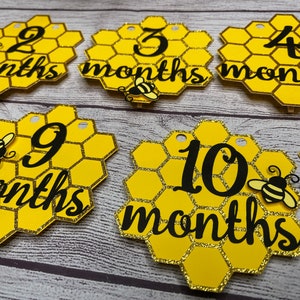 May include: Honeycomb-shaped monthly milestone markers in yellow and gold glitter. Each marker features a number from 2 to 10, with the word "months" in black script. Adorable bee cutouts are attached to some of the markers. These are perfect for baby photos.