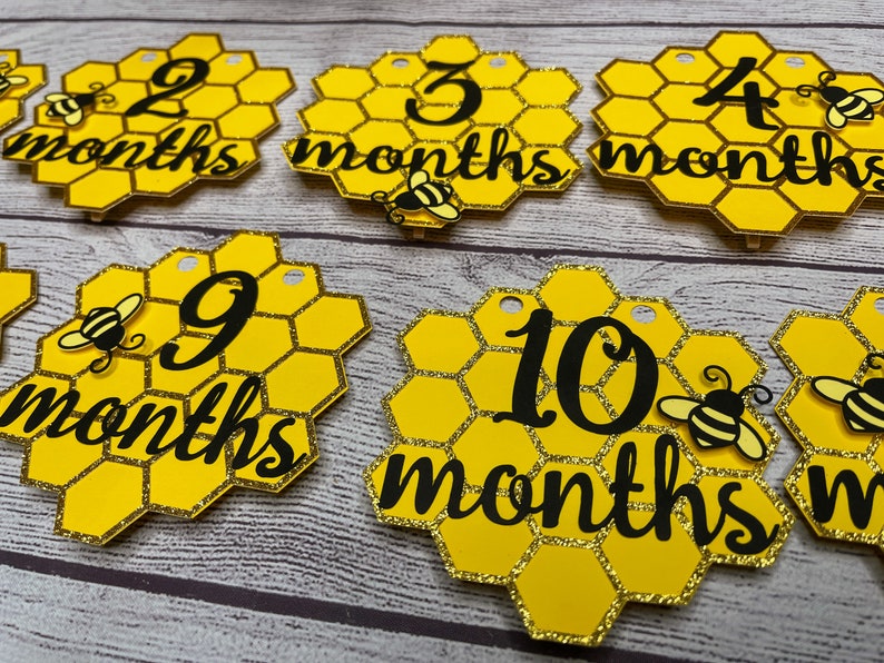 May include: Honeycomb-shaped monthly milestone markers in yellow and gold glitter. Each marker features a number from 2 to 10, with the word "months" in black script. Adorable bee cutouts are attached to some of the markers. These are perfect for baby photos.