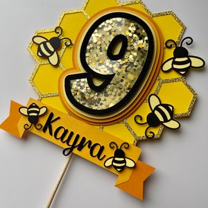 May include: A yellow and black birthday cake topper featuring the number 9, set within a honeycomb design and decorated with bee motifs. The name "Kayra" is written on a banner. The topper is attached to a wooden stick.