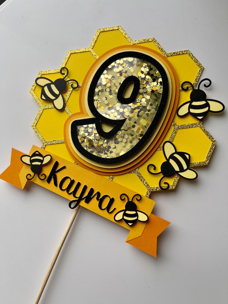 May include: A yellow and black birthday cake topper featuring the number 9, set within a honeycomb design and decorated with bee motifs. The name "Kayra" is written on a banner. The topper is attached to a wooden stick.