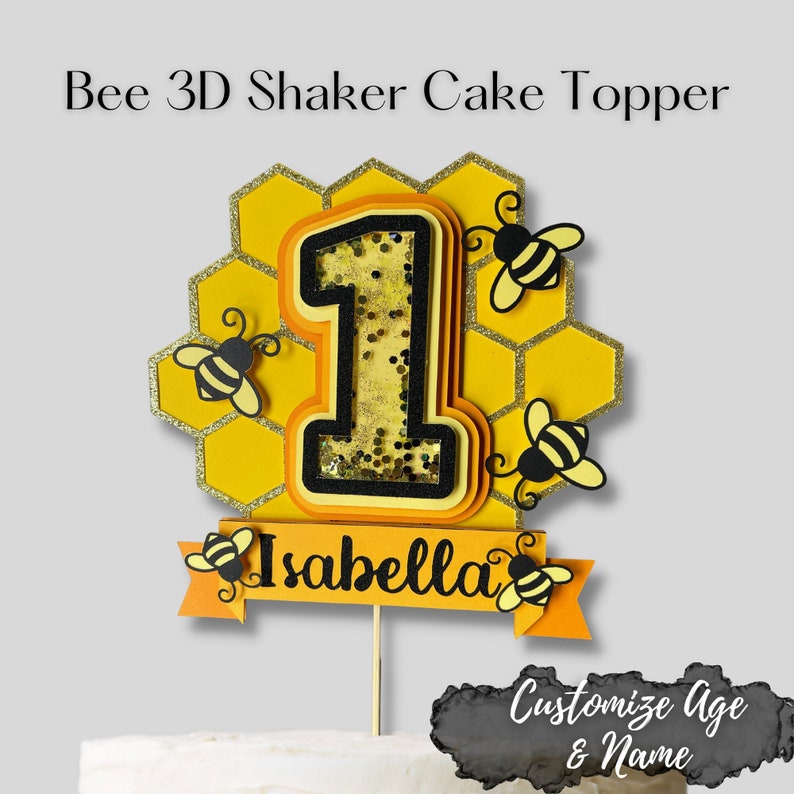 May include: A bee-themed cake topper featuring the number 1, a honeycomb design, and the name "Isabella." The number 1 is filled with gold glitter. The topper includes small bee cutouts and the text "Customize Age & Name."
