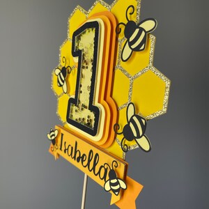 May include: A yellow and black bee-themed cake topper with the number "1" and the name "Isabella". The topper features a honeycomb design, glitter accents, and small bee cutouts. The number "1" has a shaker element.