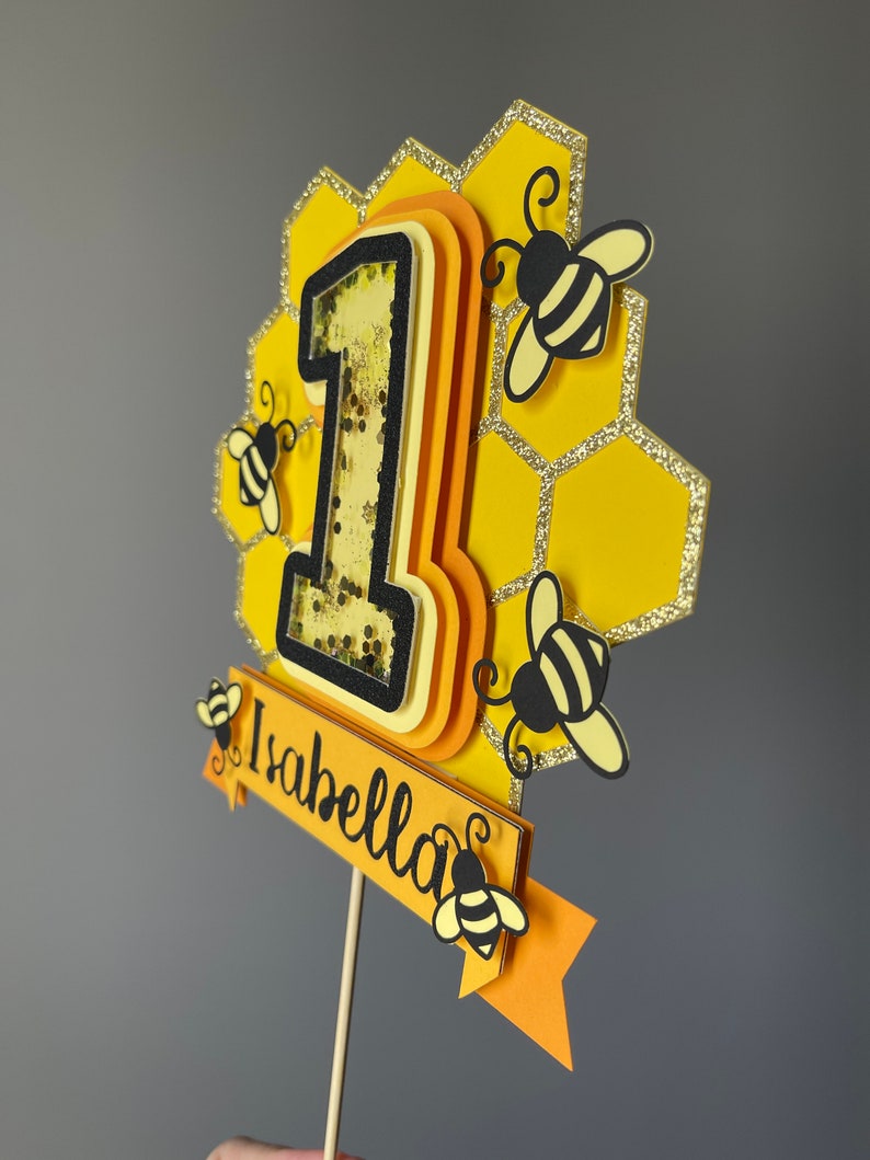 May include: A yellow and black bee-themed cake topper with the number "1" and the name "Isabella". The topper features a honeycomb design, glitter accents, and small bee cutouts. The number "1" has a shaker element.