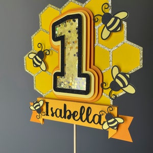 May include: A layered cake topper featuring the number one in yellow and black, with a honeycomb background and glitter details. The number one is filled with confetti. The name "Isabella" is on a banner, with bee cutouts.