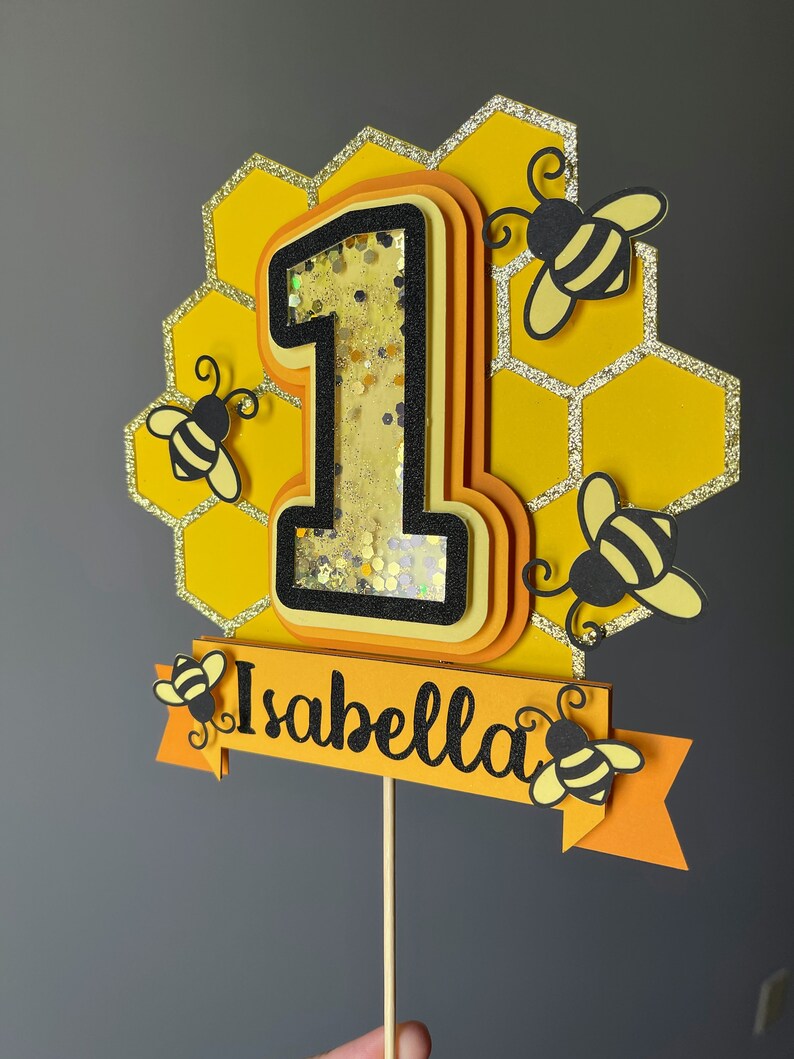 May include: A layered cake topper featuring the number one in yellow and black, with a honeycomb background and glitter details. The number one is filled with confetti. The name "Isabella" is on a banner, with bee cutouts.