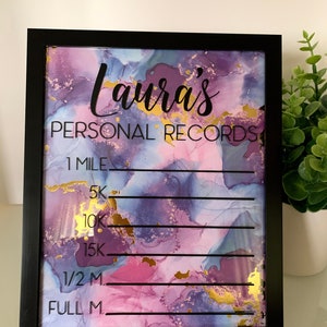 May include: A framed dry-erase board with the text "Laura's PERSONAL RECORDS" in black script. The background features a watercolor design in shades of purple, pink, and gold. The board includes lines for recording running distances such as 1 mile, 5K, and a full marathon.