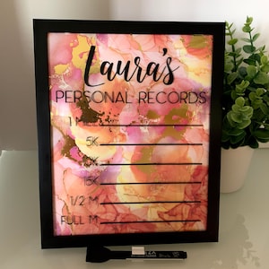 May include: A framed sign with the text "Laura's PERSONAL RECORDS" in black lettering. The background features a colorful abstract design in shades of pink, gold, and peach. Includes a dry erase marker.