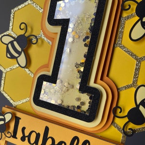 May include: A layered cake topper featuring a number one filled with glitter, and the name "Isabella" in black script. The design includes yellow and black honeycomb shapes and bee cutouts, ideal for a first birthday celebration.