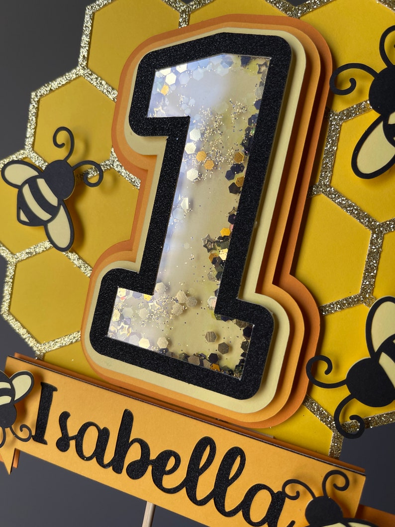 May include: A layered cake topper featuring a number one filled with glitter, and the name "Isabella" in black script. The design includes yellow and black honeycomb shapes and bee cutouts, ideal for a first birthday celebration.