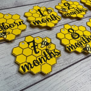 May include: Honeycomb-shaped milestone markers in yellow and gold glitter. Each marker features a black script indicating a baby's age, from "newborn" to "9 months." Adorable bee accents adorn each piece, creating a charming theme.