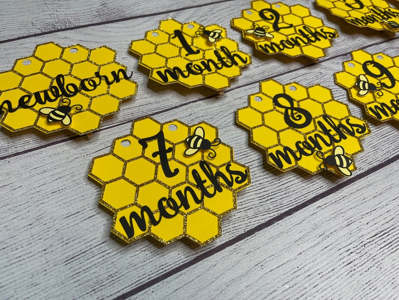 May include: Honeycomb-shaped milestone markers in yellow and gold glitter. Each marker features a black script indicating a baby's age, from "newborn" to "9 months." Adorable bee accents adorn each piece, creating a charming theme.