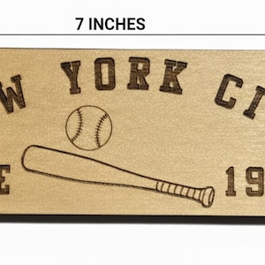 May include: A wooden bottle opener, 7 inches long and 1.5 inches wide, with a circular hole and a bottle cap opener. The text "NEW YORK CITY SINCE 1903" is engraved, along with a baseball and bat.