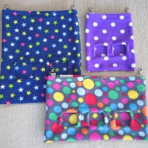 Op de afbeelding: Drie hangende fleece-dierenbedden met verschillende patronen. Het eerste bed is blauw met witte en groene sterren. Het tweede bed is paars met witte stippen. Het derde bed is grijs met kleurrijke stippen.