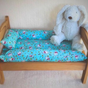 Peut inclure: Un lit de poupée en bois avec un ensemble de literie à imprimé de lapin bleu et blanc. Un lapin en peluche blanc est assis sur le lit.