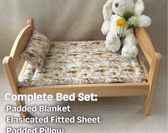 May include: A wooden doll bed with a floral patterned fitted sheet, a padded blanket, and a padded pillow. A white stuffed bunny with yellow flowers sits on the bed. The text "Complete Bed Set: Padded Blanket Elasicated Fitted Sheet Padded Pillow" is below the bed.