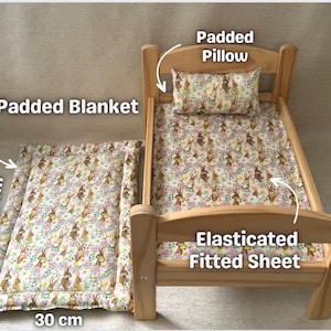 Op de afbeelding: Een houten poppenbed met een gevoerd dekbed, een hoeslaken en een kussen. De beddengoed heeft een patroon van konijnen en bloemen. Het bed meet 30 cm bij 48 cm.