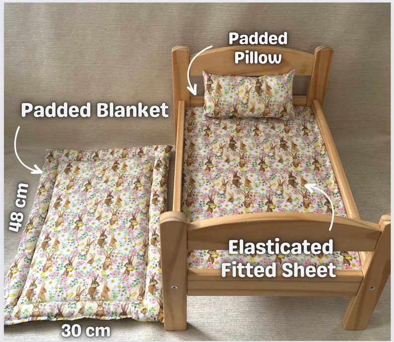 Op de afbeelding: Een houten poppenbed met een gevoerd dekbed, een hoeslaken en een kussen. De beddengoed heeft een patroon van konijnen en bloemen. Het bed meet 30 cm bij 48 cm.