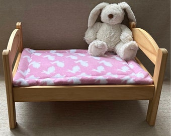 May include: A wooden doll's bed with a pink and white patterned mattress and a white plush bunny sitting on the bed.