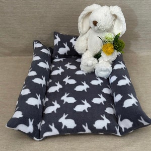 Puede incluir: Una cama para mascotas de felpa gris y blanca con un juguete de peluche de conejo blanco sentado encima. La cama tiene forma de rectángulo con esquinas redondeadas y tiene un patrón de conejo blanco.