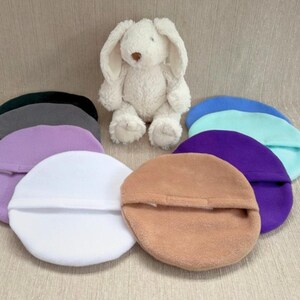 Puede incluir: Un conjunto de diez diademas redondas de felpa, cubiertas de tela, en varios colores, incluyendo blanco, morado claro, gris, azul claro, turquesa, morado y marrón. Las diademas están dispuestas en círculo alrededor de un conejito de peluche blanco.