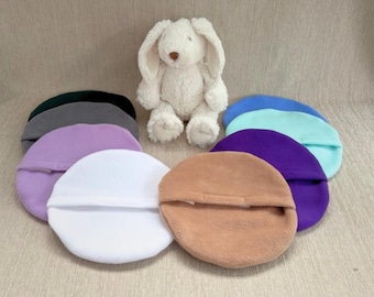 May include: A plush white rabbit toy positioned behind a collection of circular, fleece-like fabric pads in various colors including white, tan, purple, lavender, light blue, mint green, dark gray, and black, arranged in a semi-circle formation.