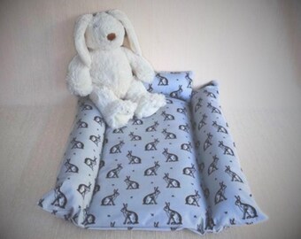 May include: A blue and white patterned baby nest with a white stuffed bunny rabbit sitting on top. The nest is made of soft fabric and has a comfortable design.