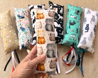 May include: A collection of six rectangular fabric catnip toys featuring various cat-themed patterns in colors including beige, white, black, and teal. Each toy is stuffed and finished with multiple colorful satin ribbons attached at the base.