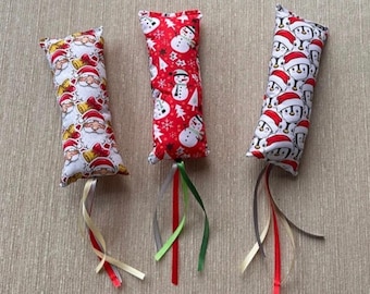 Può includere: Tre giocattoli rettangolari in tessuto per gatti con motivi natalizi. Il giocattolo a sinistra presenta Babbo Natale e campanelle su bianco, quello centrale pupazzi di neve su rosso, e quello a destra pinguini con cappelli natalizi. Nastri pendenti.