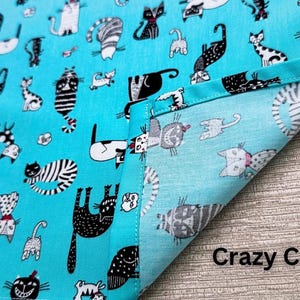 May include: A turquoise fabric with a repeating pattern of whimsical cat illustrations in black, white, and gray. The phrase "Crazy Cats" is printed in white at the bottom right corner. The fabric is folded over, showing the reverse side.
