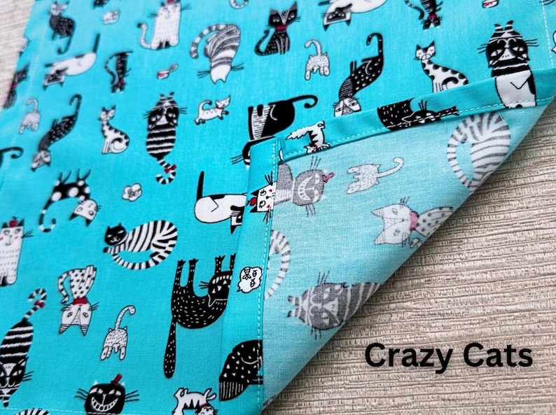 May include: A turquoise fabric with a repeating pattern of whimsical cat illustrations in black, white, and gray. The phrase "Crazy Cats" is printed in white at the bottom right corner. The fabric is folded over, showing the reverse side.