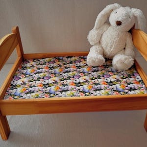 Puede incluir: Una pequeña cama de madera con un colchón estampado floral con conejitos y flores. Un conejito de peluche blanco está sentado en la cama. El marco de la cama es de madera clara.