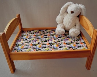 May include: A small wooden bed with a floral patterned mattress featuring bunnies and flowers. A white plush bunny sits on the bed. The bed frame is made of light-colored wood.