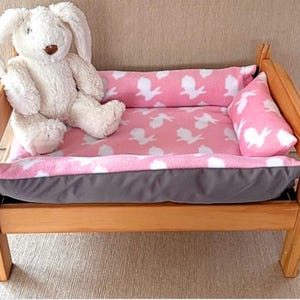 May include: A small wooden pet bed with a pink and white bunny print cushion and pillow. A white stuffed bunny sits on the bed. The bed has a grey bottom cushion.