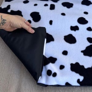 Op de afbeelding: Een deken met koeienprint, met zwarte vlekken op een witte achtergrond. De achterkant van de deken is zwart. Een hoek is omgeslagen en toont het contrast van de materialen. De deken is waarschijnlijk gemaakt van een zachte stof.